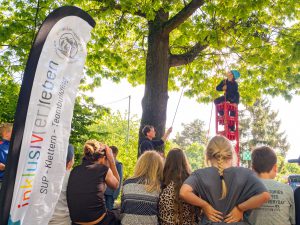 Die Menschen beobachten, wie ein mit einem Klettergurt gesicherter Kletterer im Freien in der Nähe eines „inklusiv erleben“-Banners rote Kisten erklimmt. Foto: Martin Kirchner