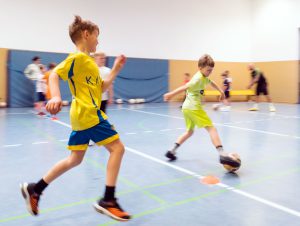 Zwei Jungen in Sportkleidung jagen in einer Sporthalle einem Fußball hinterher; im Hintergrund sind weitere Kinder zu sehen. Foto: Martin Kirchner