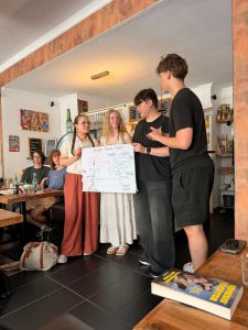 Vier junge Leute in einem Café zeigen den Gästen an den benachbarten Tischen ein handgeschriebenes Plakat. Foto: Evangelisches Gymnasium Siegen-Weidenau