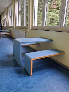 Minimalistische blaue Bank mit Tisch vor großen Fenstern in einem Flur; draußen sind Bäume zu sehen.