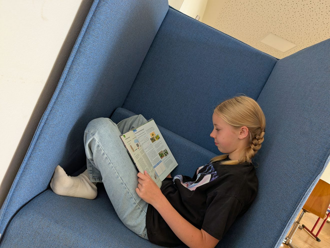 Ein blondes Mädchen mit Zöpfen liest auf einem blauen Sofa mit hohen Lehnen eine Zeitschrift. Foto: Evangelische Werkschule Naundorf