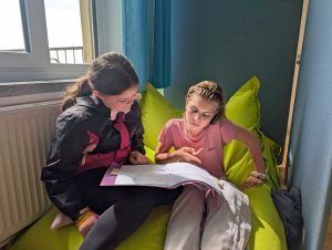 Zwei Mädchen sitzen auf grünen Sitzsäcken vor einem sonnigen Fenster, lesen und unterhalten sich gemeinsam über ein Buch. Foto: Evangelische Werkschule Naundorf