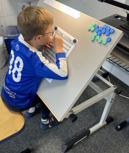 Ein Kind mit Brille schreibt an einem geneigten Schreibtisch mit blauen und grünen Scheiben und wirkt dabei ganz konzentriert. Foto: FES Böblingen