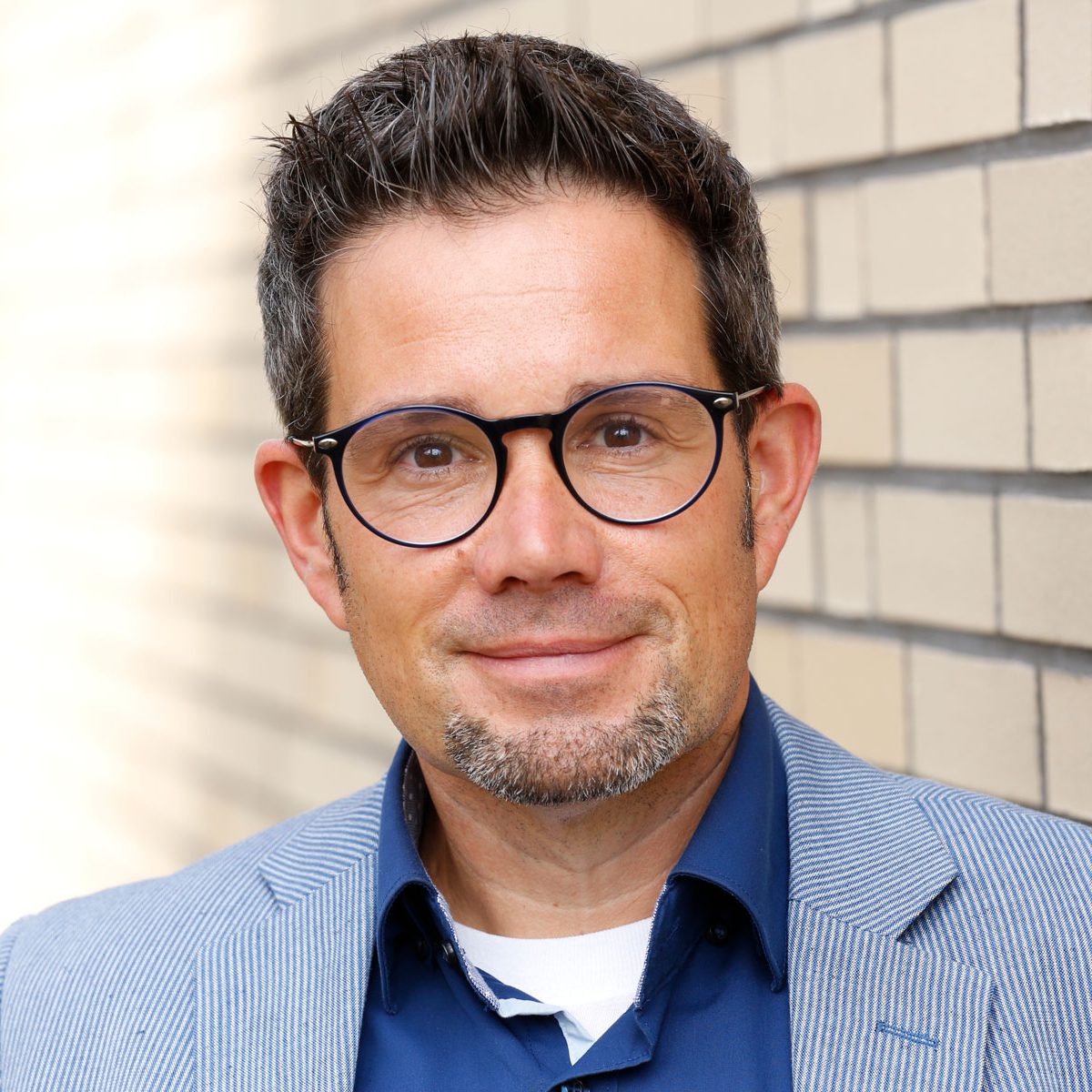 Ein Mann mit Brille und Spitzbart, der ein blaues Hemd und einen Blazer trägt, steht lächelnd vor einer Backsteinmauer.