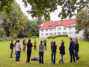 Menschen, die auf einer Rasenfläche vor einem Haus im Kreis stehen. Bild: Martin Kirchner